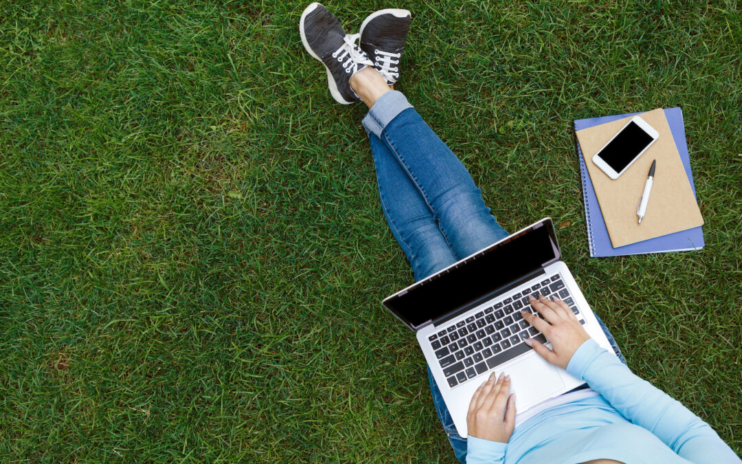 Top view of woman sitting in park on the green grass with laptop, notebook and smartphone, hands on keyboard. Student studying outdoors. Kesätyö 2024